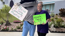 A person inside of a blue and gray stingray mascot costume stands holding a white sign, while the other arm is around the shoulder of a woman wearing a purple shirt and black leggings, holding a green sign. Both signs note a community meeting taking place on Oct. 12.