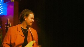 A musician in an orange jacket plays a white electric guitar on a dimly lit stage.
