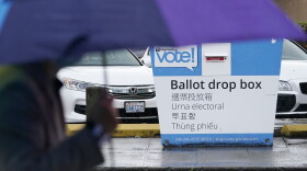 A person carrying an umbrella walks past a King County Ballot Drop box with cars parked behind it.