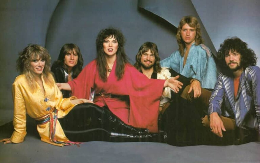 Six band members - two women and four men - sitting on the floor and looking into the camera. All have long hair