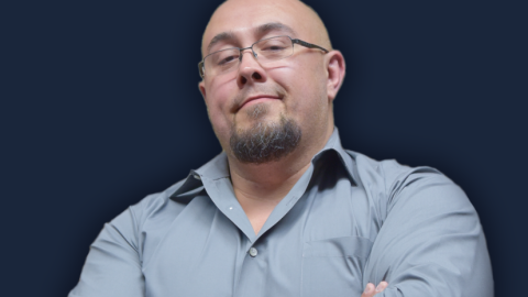 A man in a light blue shirt crosses his arms in front of a navy background. 