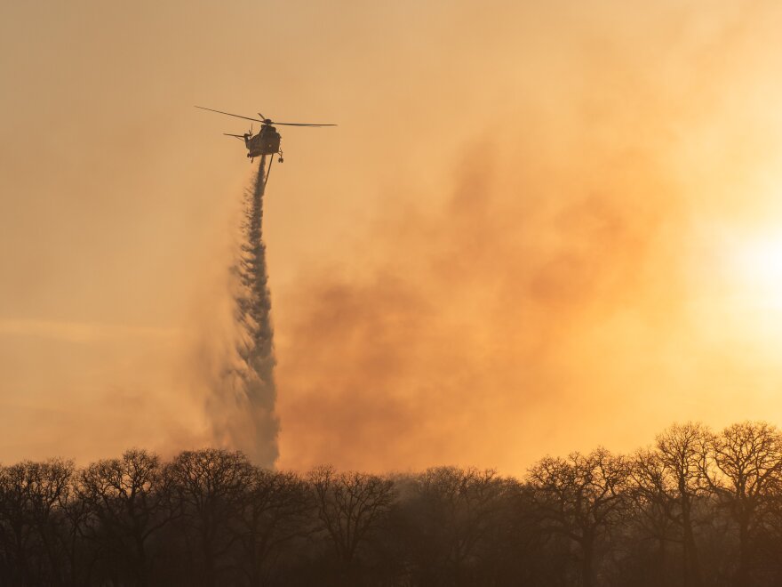 A helicopter drops fire retardant over a forested area as smoke rises in the background