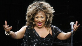 Tina Turner wearing a black dress with sequins with arms stretched outward.