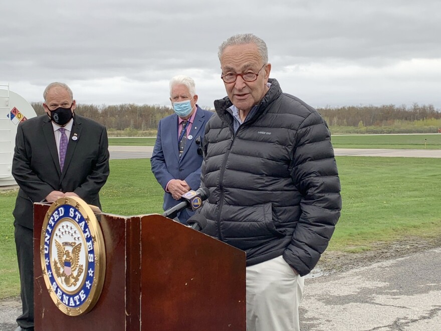 Senator Chuck Schumer stands at a podium. He is wearing glasses, has grey hair, and is wearing a black coat and tan pants. There are two men wearing face masks and suits in the background behind him.