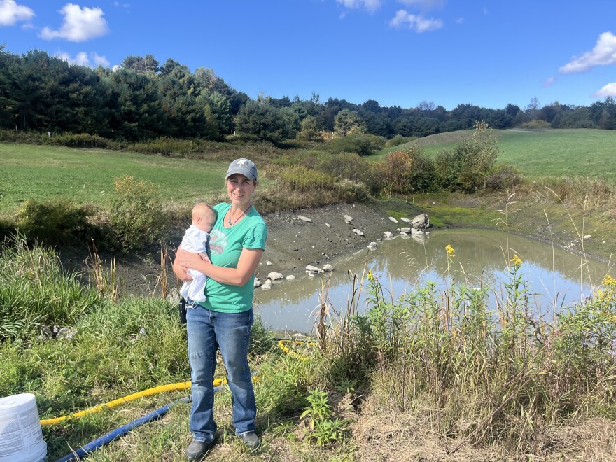 A woman and her baby stand in front of a pond.
