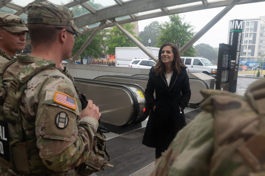 A woman in a black overcoat talks with soldiers in camouflage uniforms in a subway station. 