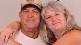 Steven and Gina Poole pose together at their StoryCorps appointment in Saint George in 2025. Steven wears a tan baseball cap with a cactus A.T.T.I. logo on it, and a white tee shirt with a picture of a brick wall and a hooded person standing in front of it. The text above and below the is partially obscured by Gina's arm.  What can be seen reads "be an organ don if the alley is dark enough." Gina stands facing the camera with her arms around Steven.  She wears glasses pushed up onto her head. Her long gray hair has bangs.  She wears a maroon sleevlelss tee.  They are both smiling with closed lips.