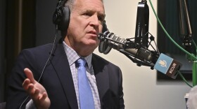A man sitting inside a radio studio talks at a microphone. he is gesturing with his right hand.