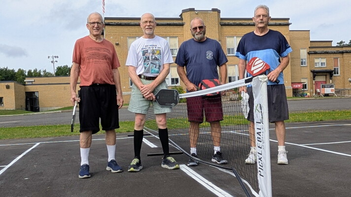 Men stand on both sides of a pickleball net in front of an old school building.