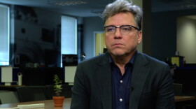 A man with a black blazer and glasses frowning but just a little bit
