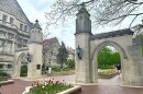 The Sample Gates at Indiana University Bloomington. They are limestone arches with black sconces. Red and white tulips are planted nearby. Students walk in the background.