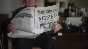 An older man sits in a desk chair and holds the Marion County Record newspaper published after the outlet was raided by local police. The headline on the front page, in large text, reads "SEIZED...but not silenced." There's a story on the right side of the front page that has the headline "KBI takes over," with a one-column line of text. Next to that text is a four-column image of the police raiding the newspaper office. On the back page of the paper is a single, wide column of text, with a large quote above it reading, "Freedom of the press is as important to democracy, as democracy."