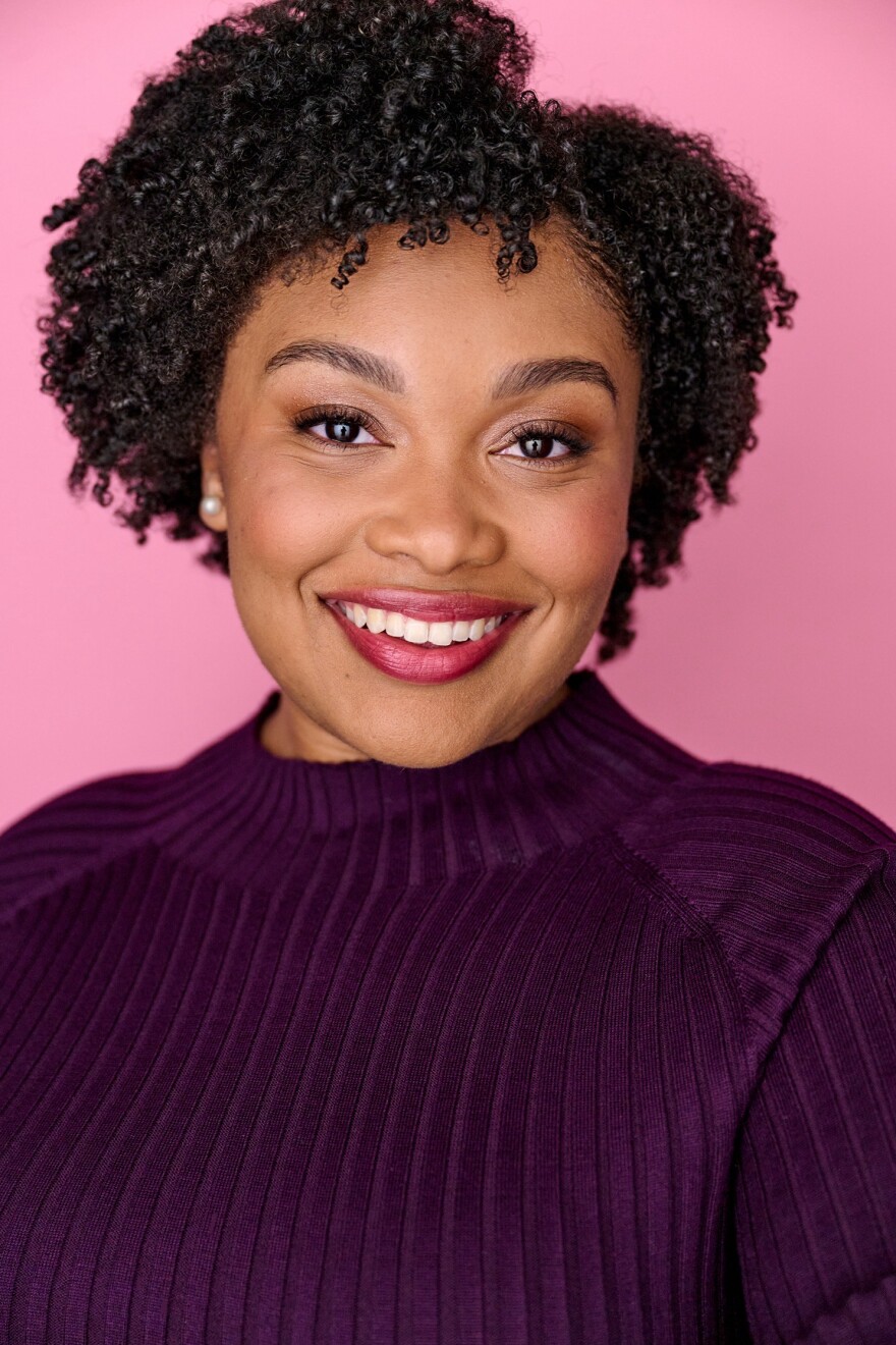 A woman wearing a purple turtleneck is smiling at the camera. She is wearing pearl stud earrings, and is standing in front of a pink background.