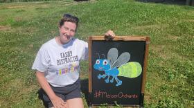 A woman kneels down in the grass at an orchard next to a chalkboard with a drawing of a cartoonish firefly