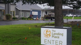 A medium sized white sign framed by a black post peeks out at the bottom right of the frame. The sign reads "The Landing Place" with a stylized image of a yellow bird resting on a branch. Behind the sign sits a large tree, whose branches cover the top of the frame. Behind both of those subjects is the Landing Place, a gray building with one segment in the middle of the image painted blue. Landscapers can be seen working the exterior of the building. The weather is overcast, with sporadic puddles of water seen in the Landing Places' parking lot. 