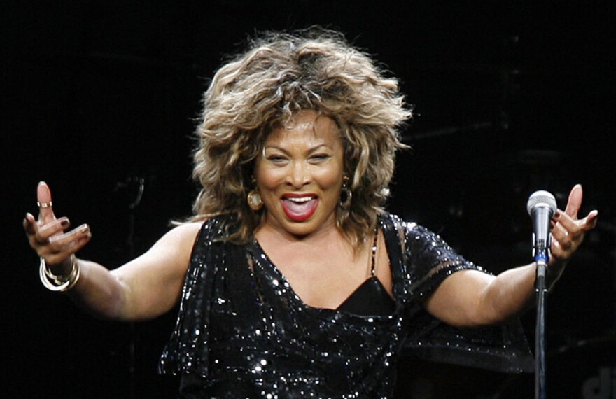 Tina Turner wearing a black dress with sequins with arms stretched outward.