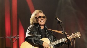 Medium shot of an older white male with light colored hair past his collar, wearing sunglasses, a black shirt of shiny fabric holding an acoustic guitar and standing in front of a microphone at an indoor venue.