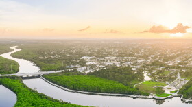 Aerial view at sunrise or sunset of a river winding through dense green mangroves, with a bridge crossing the water and a long boardwalk along the shoreline. A park with lawns, walking paths and a parking lot sits at right, with neighborhoods stretching toward a hazy horizon.