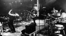 A black-and-white image of four men playing instruments on stage in a theater with a crowd of people watching.