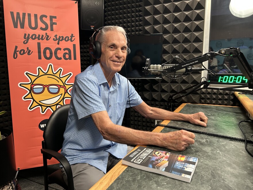 Older man sits in front of microphone with book on studio desk with WUSF Your Spot for Local sign. 