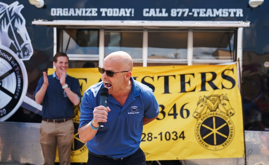 State Rep. Dave Lislegard's mouth is wide open as he yells into a microphone. He is standing in front of a trailer with Teamsters logos.