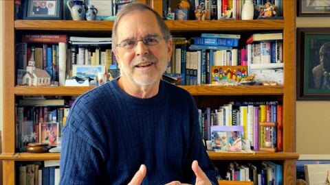 Speaking in front of a bookcase, a man smiles and weaves his fingers together