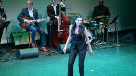 Woman with long brown hair tap dances on stage with band behind her 