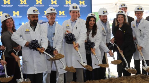 A group of people are lined up wearing white lab coats and hard hats with University of Michigan branding. They are all smiling while holding shovels, digging up a mound of dirt to symbolically break ground for the construction project.