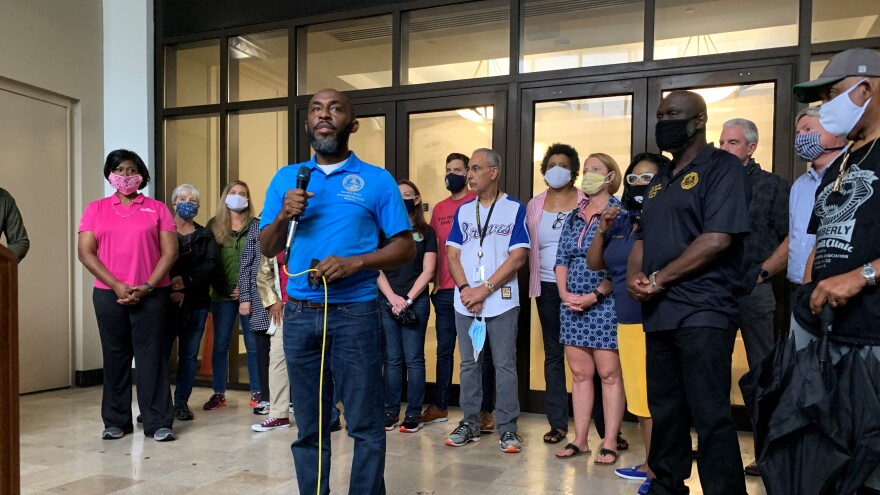 Garrett Dennis speakign with a mic in his hand, with other council members and Florida politicans behind him, right in front of the entrance of City Hall.