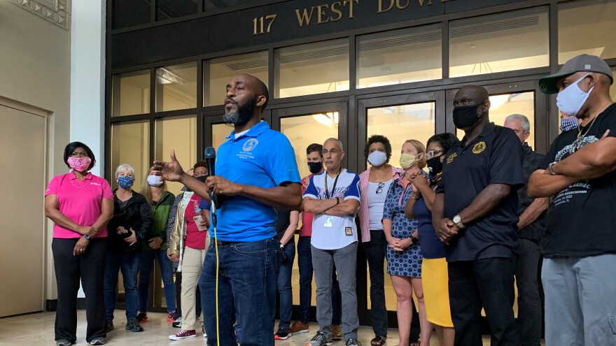 Dennis speaking with a mic in his hand in front of the doors of city hall, right in front of the doors are a bunch of elected officials, including city council people and state officials.