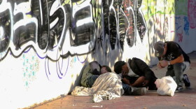 Three people slumped over on a sidewalk near a wall covered in graffiti.