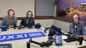 Three smiling men wearing headphones sit at a table in a radio talk studio: a man at left has short brown hair and is wearing a blue and white plaid button-down shirt; a man at center has short black hair and is wearing glasses and a black shirt under a dark grey suit coat; a man at right has very short dark hair and is wearing glasses and a blue button-down shirt