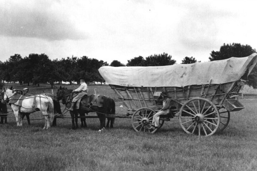 A black and white photo of a Conestoga wagon in a field, pulled by a team of horses. Two men are visible, one riding a horse and the other seated on the front of the wagon. The wagon has a large canvas cover and is a type of heavy, covered wagon used in the 18th and 19th centuries. 