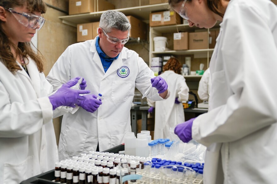 Several people in white lab coats and wearing purple gloves and goggles look down at containers holding samples for testing