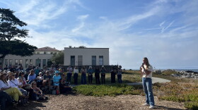 A woman stands holding a microphone with seated and standing audience members looking at her on the edge of the coast. 