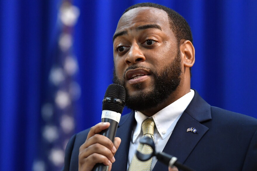 a black man in a suit and tie with a flag pin on his label holds a microphone