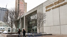 Outdoors photo showing the exterior of a large concrete building with trees in the front and two people walking in the foreground. A sign on the building reads: "Crown Center, shops and restaurants."