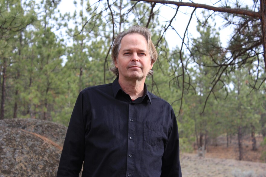 A man wearing a black button down shirt stands in the woods.