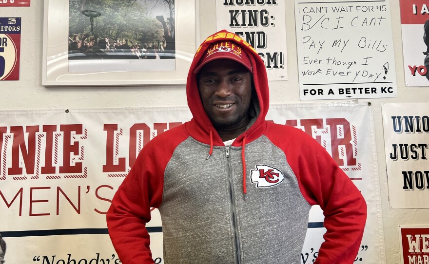 A man in a red and grey hoodie with the Chiefs logo stands with his hands on his hips. He's in front of a wall with posters that demand a living wage.