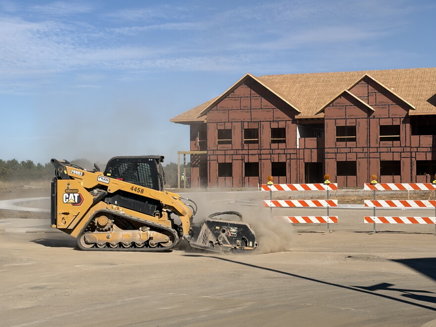 A yellow bulldozer uses a brush attachment to sand down poured concrete in front of a framed condo structure 