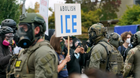 Anti-ICE protestors stand facing police officers who are wearing gas masks and camouflage fatigues.