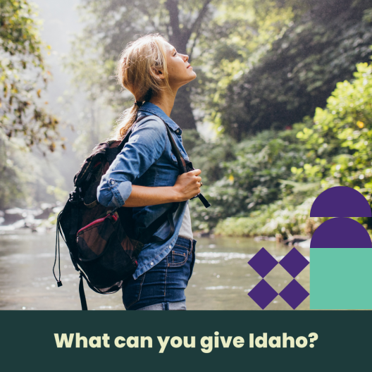 A woman wearing a denim shirt and shorts with a backpack, standing in a body of water with the words 'what can you give Idaho' at the bottom. 