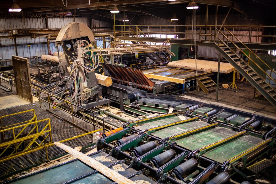 The inside of a sawmill. A pre-cut log sticks out of a machine on its path to becoming planks of lumber.