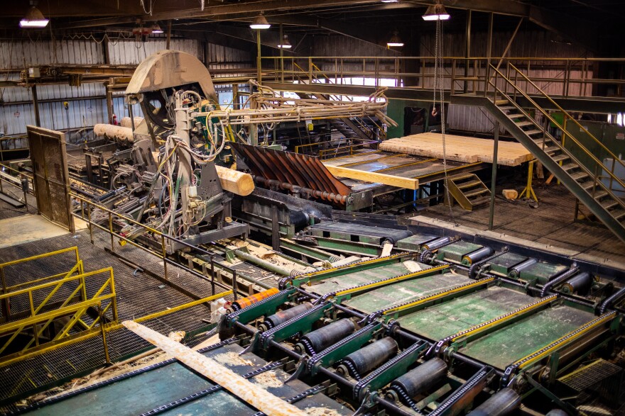 The inside of a sawmill. A pre-cut log sticks out of a machine on its path to becoming planks of lumber.