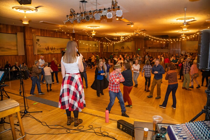 Ava Honey calls square dancing on stage with a microphone in hand, looking out at a floor full of people dancing.