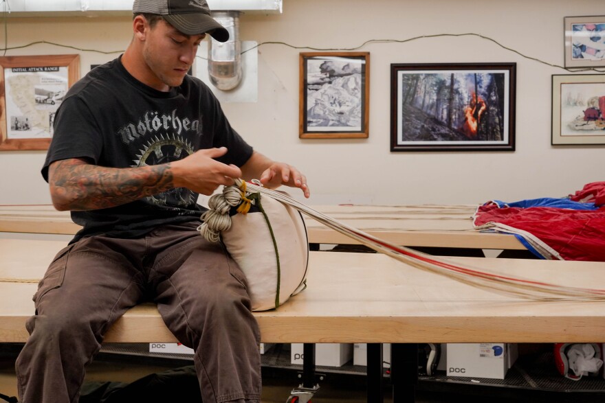 A man sits on a table and packs a parachute. There is a picture of a wildfire on the wall behind him.