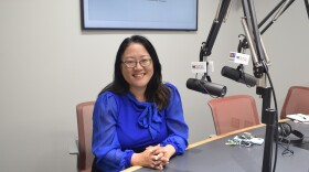Woman in blue top smiling as she sets in a radio studio next to a microphone and a monitor that reads 'The Vidette' behind her