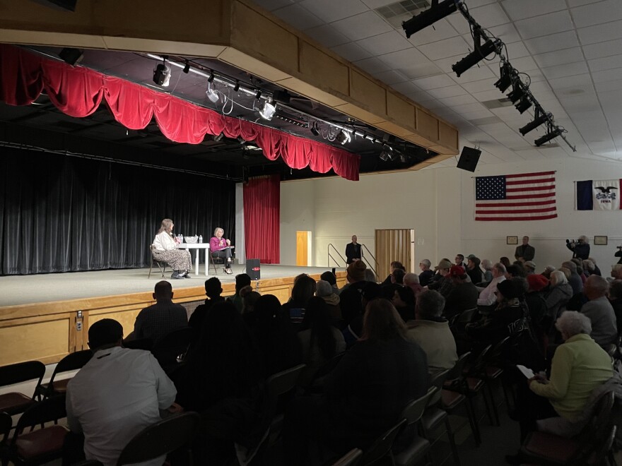 Two women sit on a stage in front of a large crowd in an auditorium