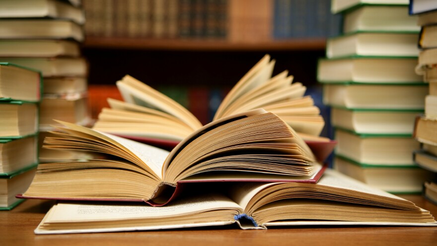 Stacks of books are on a table. In the center, three books are open to various pages.