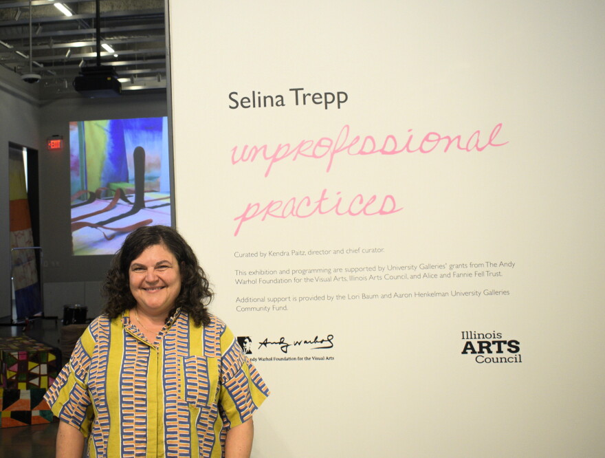 A woman smiles while standing next to a wall displaying text that reads “Selina Trepp unprofessional practices” at an art exhibit. Bright art is visible in the background.